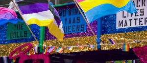 A vibrant Pride float decorated with gold and blue glitter features signs reading “THE DOLLS,” “NO KINGS YAAS QUEENS,” and “BLACK LIVES MATTER.” Flags for the bisexual, nonbinary, pansexual, and other identities wave above, alongside strings of rainbow bunting.