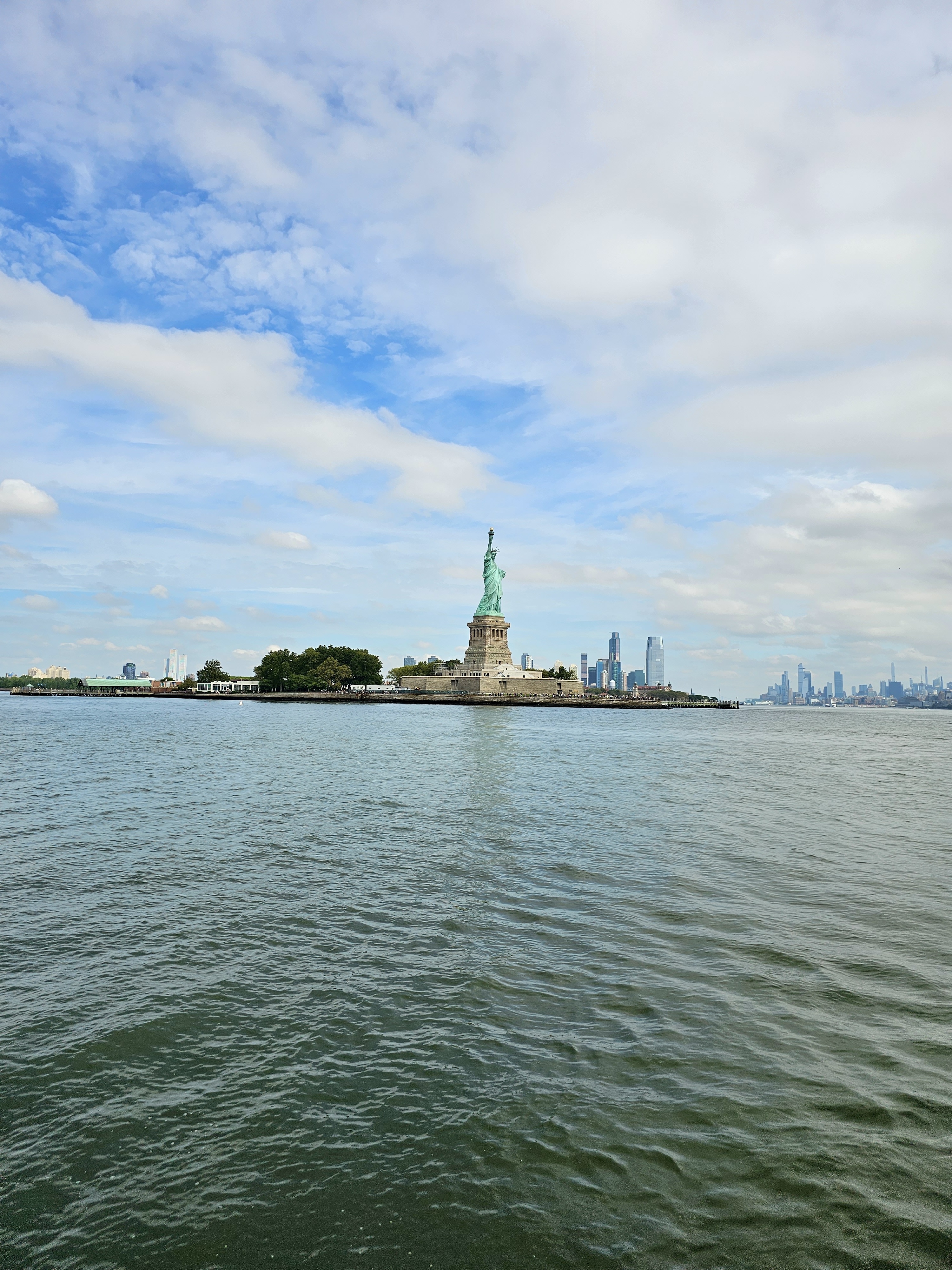 The Statue of Liberty stands tall on Liberty Island, with the NYC skyline in the background under a partly cloudy sky.