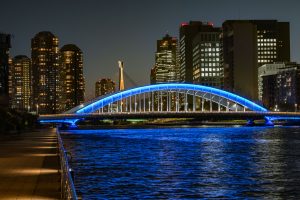 The Eitai Bridge (Koto Ward, Tokyo) over the Sumida River is lit up in blue at night, and the surface of the Sumida River also shimmers blue. In the background, the high-rise apartment buildings of Tsukishima stand tall.