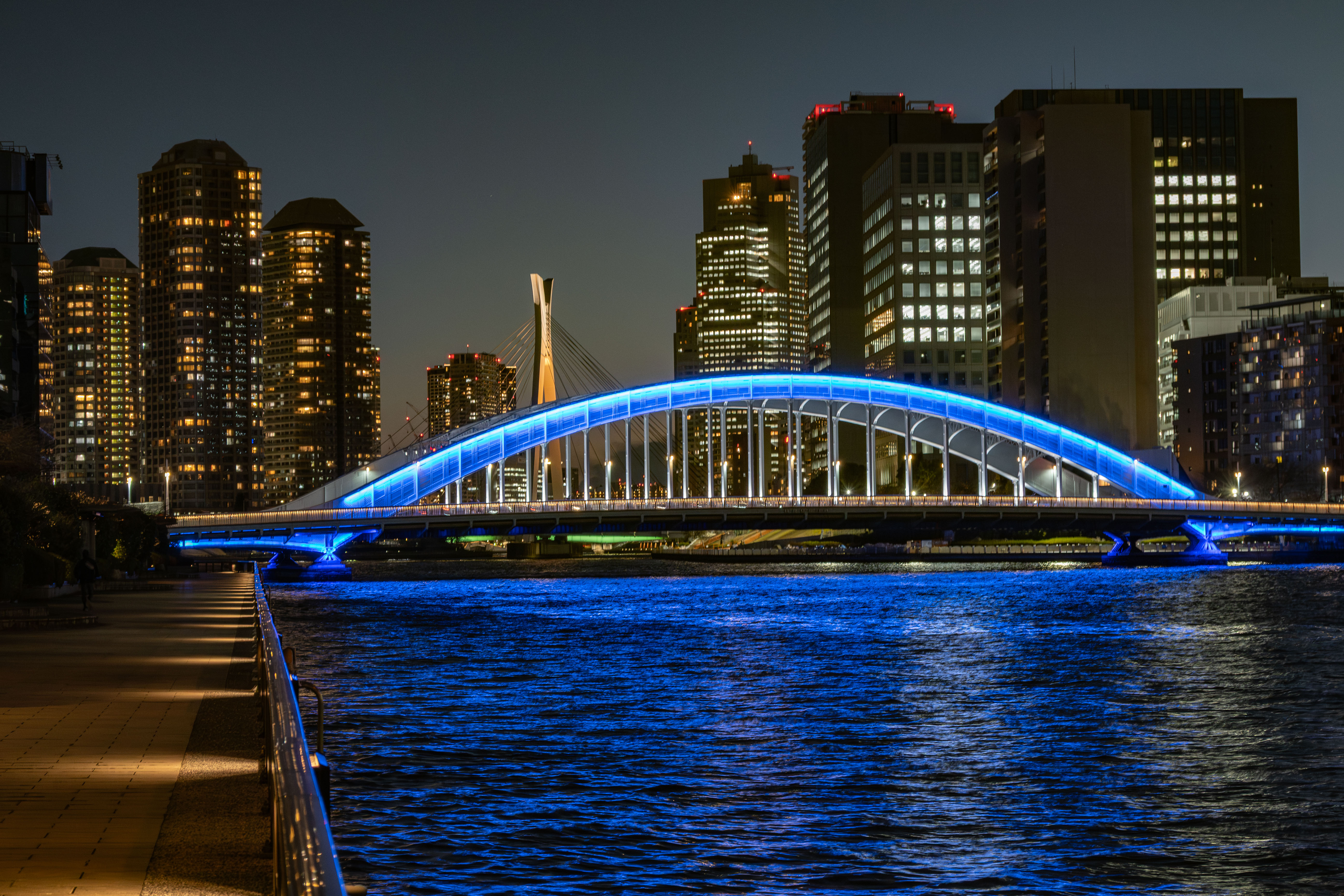 The Eitai Bridge (Koto Ward, Tokyo) over the Sumida River is lit up in blue at night, and the surface of the Sumida River also shimmers blue. In the background, the high-rise apartment buildings of Tsukishima stand tall.