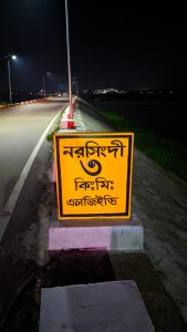 A road at night, lit by street lamps. In the foreground is a very prominent road sign.