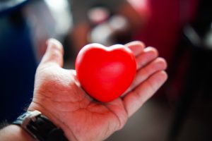 A hand holding a bright red, heart-shaped stress ball against a blurred background.
