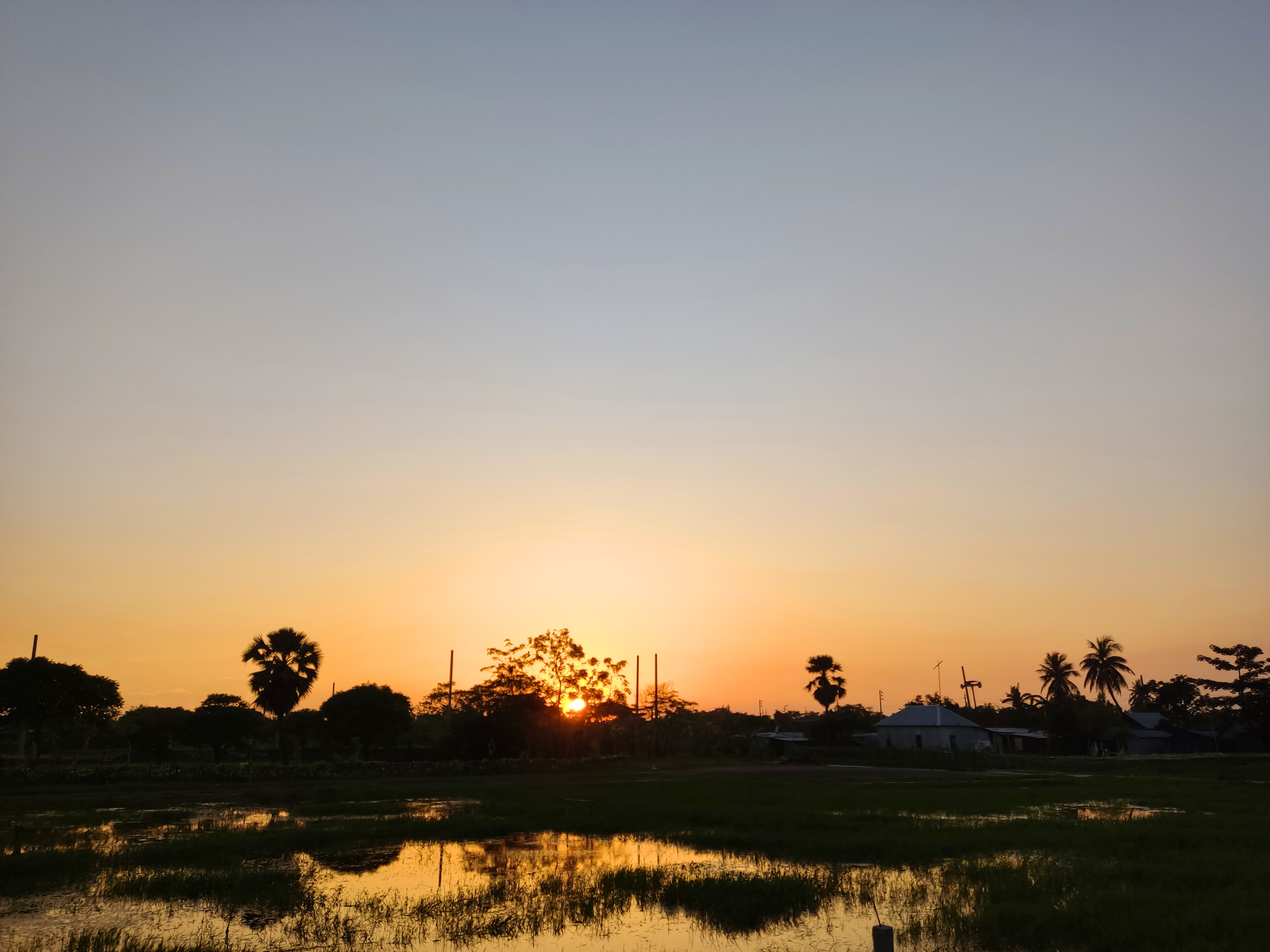 A serene sunset scene over a rural landscape, featuring a glowing orange sun dipping below the horizon.