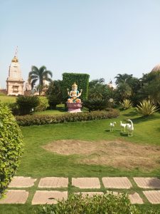 A well-maintained garden with lush green grass, decorative plants, and a stone pathway in the foreground. At the center stands a colorful statue of a Hindu deity seated on a lotus pedestal. To the right, there are three white decorative duck statues placed on the grass. 