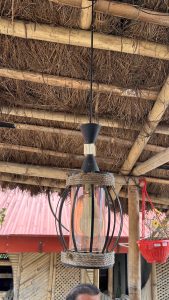 A hanging lantern with a black metal frame and a clear glass bulb, suspended from a straw-thatch roof supported by wooden beams.
