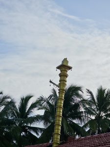 A tall, golden Dwajasthambam (temple flagpole) standing majestically against a backdrop of swaying coconut palms and a serene sky. A traditional feature seen in Kerala temples, photographed in Kozhikode.