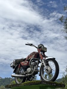 A vintage motorcycle sits atop a moss-covered platform under a partly cloudy sky.