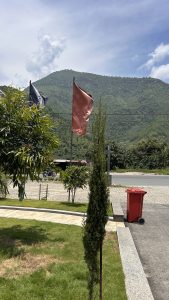 The image shows two flags of different colours, fluttering in the breeze against a backdrop of lush green mountains under a partly cloudy sky.