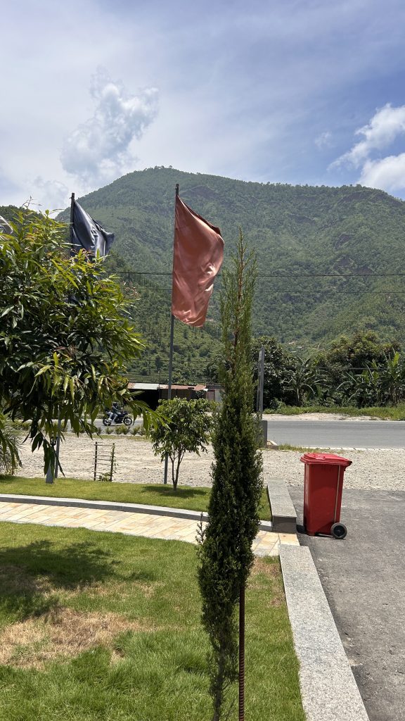 The image shows two flags of different colours, fluttering in the breeze against a backdrop of lush green mountains under a partly cloudy sky.
