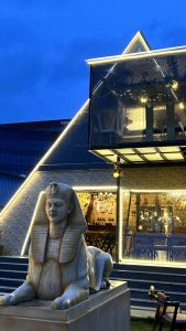 The picture of a cafe representing ancient Egypt/ Egyptian themed cafe. It's walls are decorated and painted in egyptian pattern, soft golden brown lights, wooden tables and a big egyptian statue at its front. The picture also contains clear view of night blue sky.