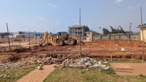 A construction site featuring a large yellow backhoe digging into brown earth. In the background, there are several buildings under construction with scaffolding. 
