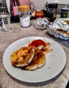 
Two golden pupusas with veggies and red sauce on a white plate. Background shows condiments and water. Pupusas, El Salvador’s national dish.
