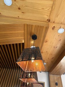 A close-up view of a modern ceiling light fixture featuring three lampshades with intricate cut-out designs of trees, casting warm light. 
