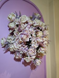 A decorative arrangement of artificial flowers, featuring a variety of roses in soft shades of cream and lavender, accompanied by small purple florals and greenery.