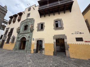 Front view of Casa de Colón in Las Palmas, featuring a carved green stone portal, wooden balconies, and a two-tone façade overlooking a cobblestone plaza.
