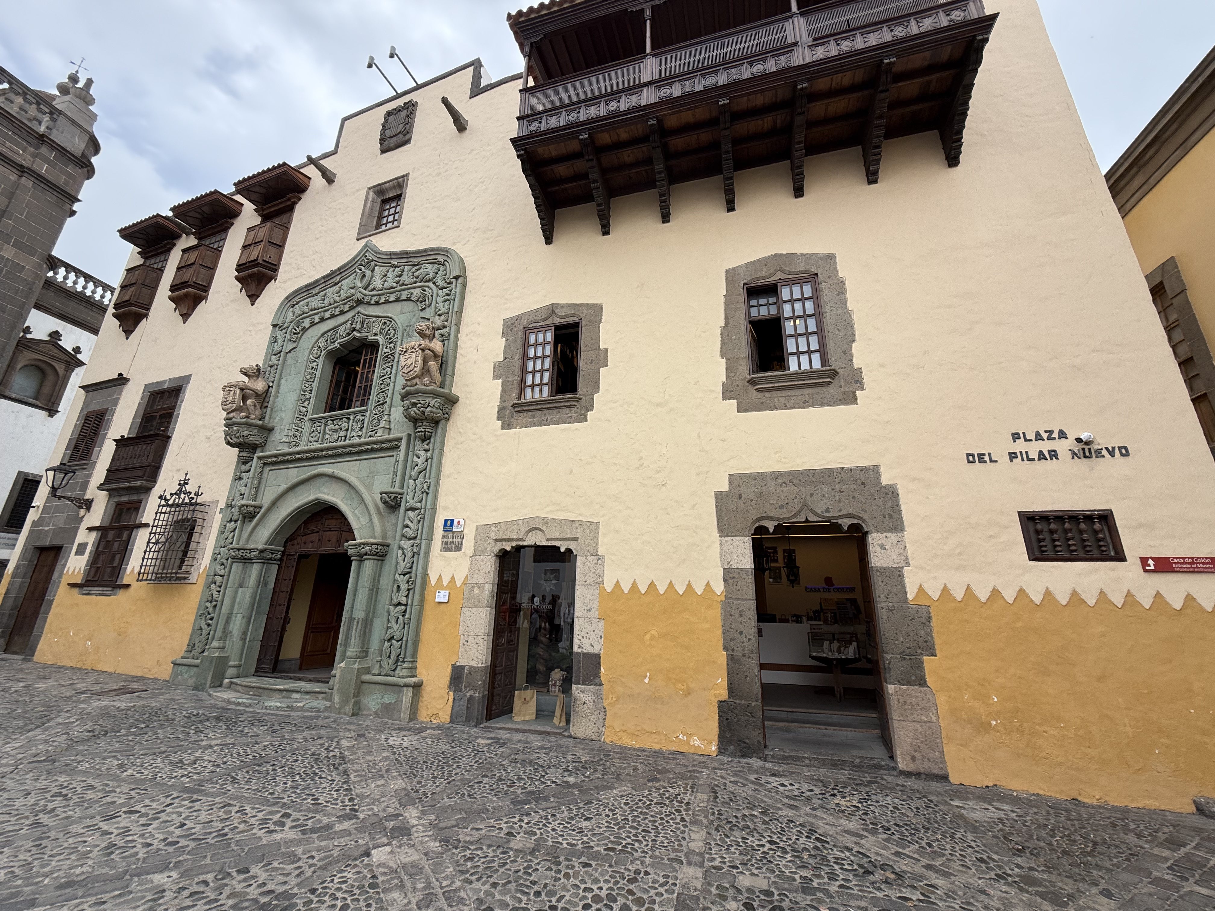 Front view of Casa de Colón in Las Palmas, featuring a carved green stone portal, wooden balconies, and a two-tone façade overlooking a cobblestone plaza.