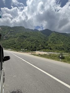 A scenic view from inside a vehicle, showcasing a winding road alongside lush, green mountains under a partly cloudy sky. 