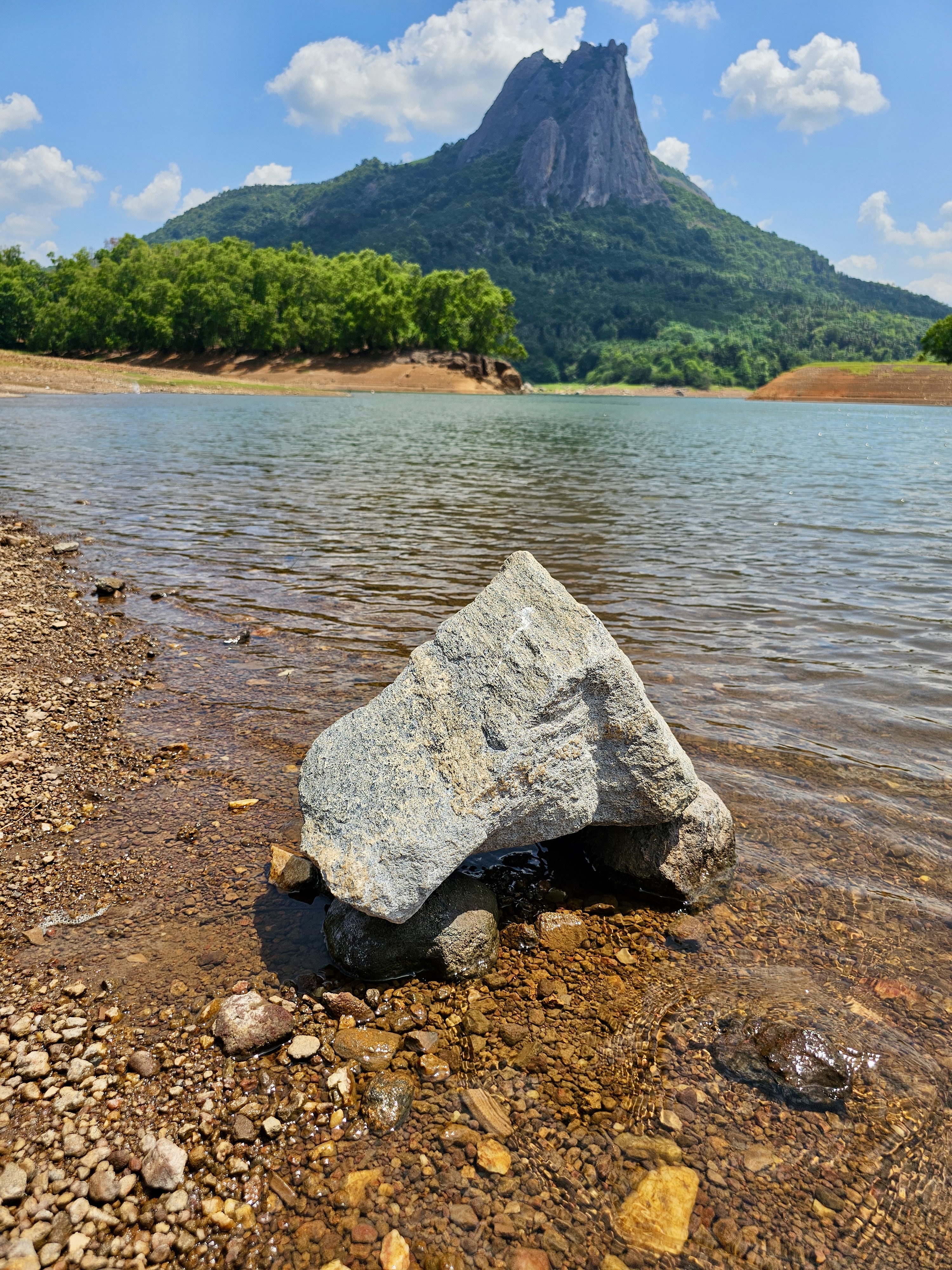 A calm and quiet view from Kanjirapuzha Dam in Palakkad, Kerala. A small rock sits in the shallow water near the edge. In the background, a tall, rough mountain rises under a blue sky with a few clouds.