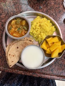 A stainless steel plate holds a variety of Indian dishes. On the plate, there is a serving of yellow rice, a bowl of mixed vegetable curry, three whole wheat rotis, a bowl of plain yogurt, and a small portion of diced mangoes. 