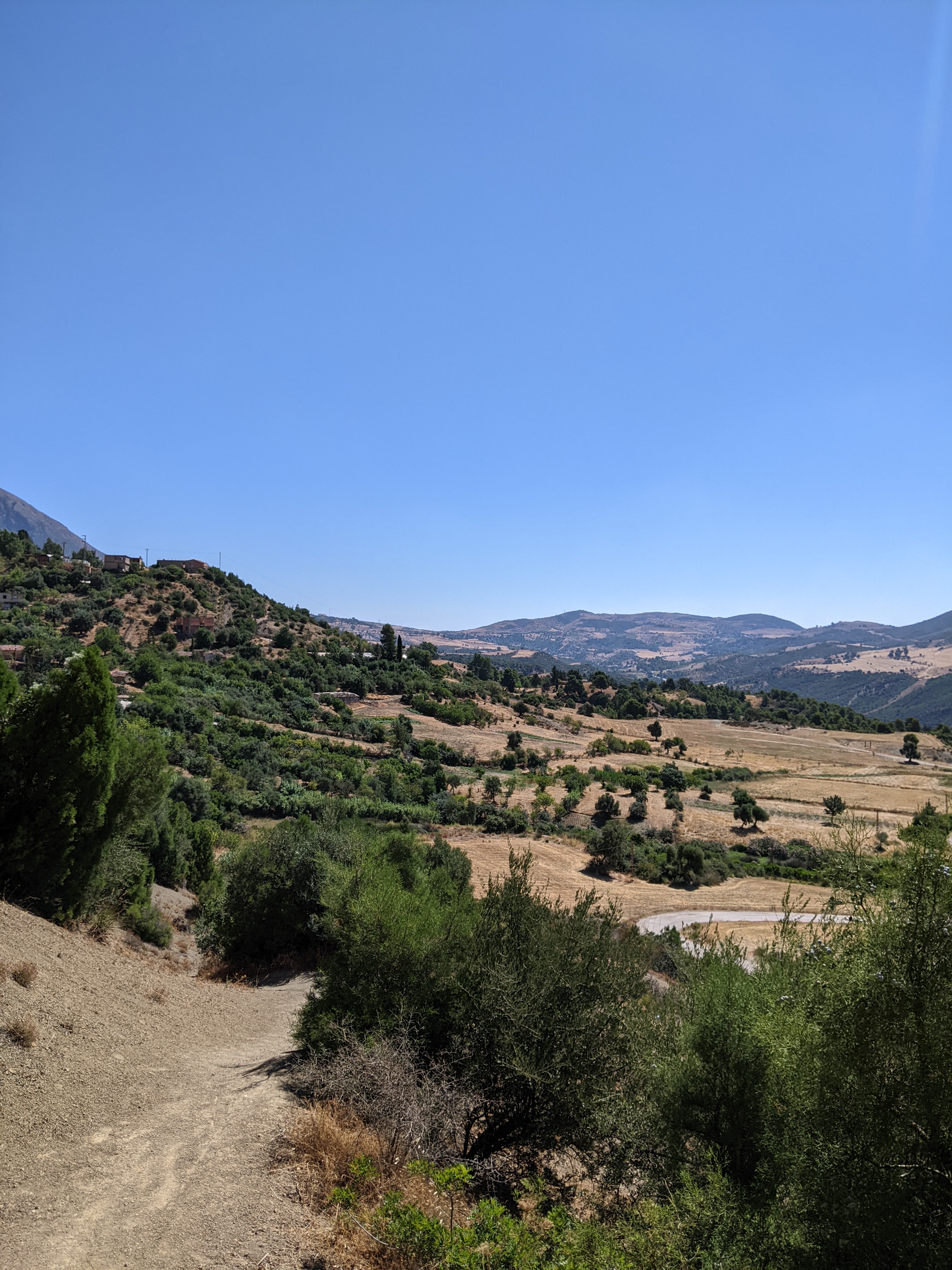 A sweeping landscape view under a clear, bright blue sky, showing rolling hills covered in a mix of green shrubs and dry, golden fields. A winding path or road is visible in the midground, and in the distance, layers of mountains fade into the haze. Some buildings can be seen on a distant hillside to the left.