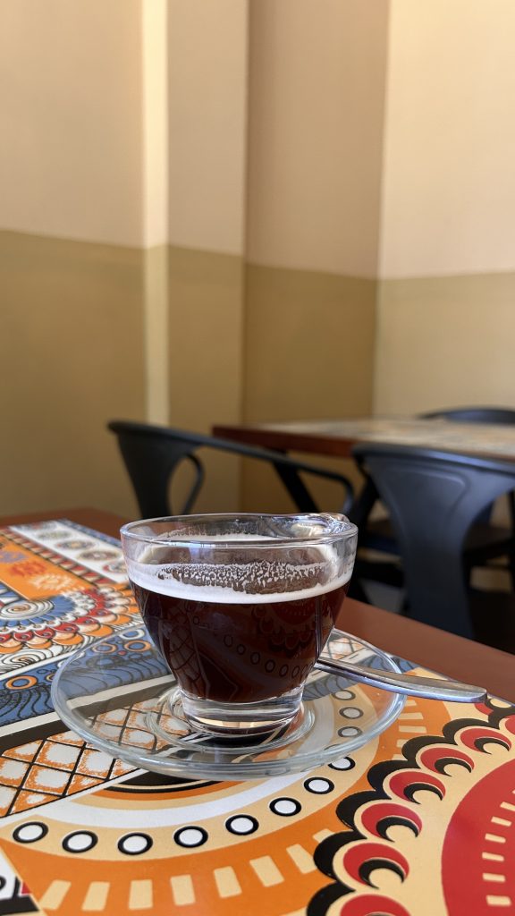 A close-up of a glass of dark coffee placed on a circular glass saucer, sitting on a colorful, patterned tablecloth.