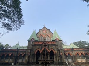 Ornate dark stone building with green spires and arched windows, framed by trees and a central fountain under a clear blue sky.
