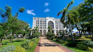 Large white multi-story building with a grand arched entrance and statues, set behind a landscaped garden with palm trees and a brick walkway.
