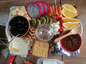 A colorful platter featuring a variety of snacks and fruits. It includes cubes of cheese, slices of dragon fruit, kiwis, carrots, cherry tomatoes, and orange segments. There are also black olives in a small bowl, mixed nuts and pretzels, cream cheese, and crackers.

