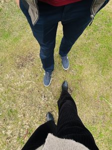 The image of a male and female standing side by side on the the green grass ground. The women is wearing a black pant and black boot while a man is wearing a blue pant and blue shoe. 