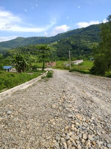 A gravel road leads through a rural landscape, flanked by lush green hills and trees under a partly cloudy sky. 