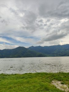 A serene landscape featuring a calm lake surrounded by lush green hills and mountains under a cloudy sky. 