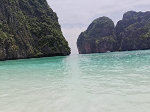 An image of a Maya Bay Island. The bay’s water is clear and green in colour, some boats can be visible. 
