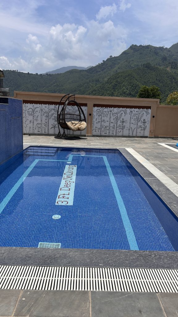 A swimming pool with blue tiled water reflecting the sky, featuring a decorative tile that reads “3 ft Deep.” To the side, there is a modern swing chair with a cushion, and in the background, a mural depicting trees and animals adorns a wall