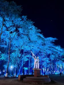 A statue of two figures holding a flag stands on a circular pedestal surrounded by trees illuminated with blue lights at night. The background features more illuminated trees and faint silhouettes of people.
