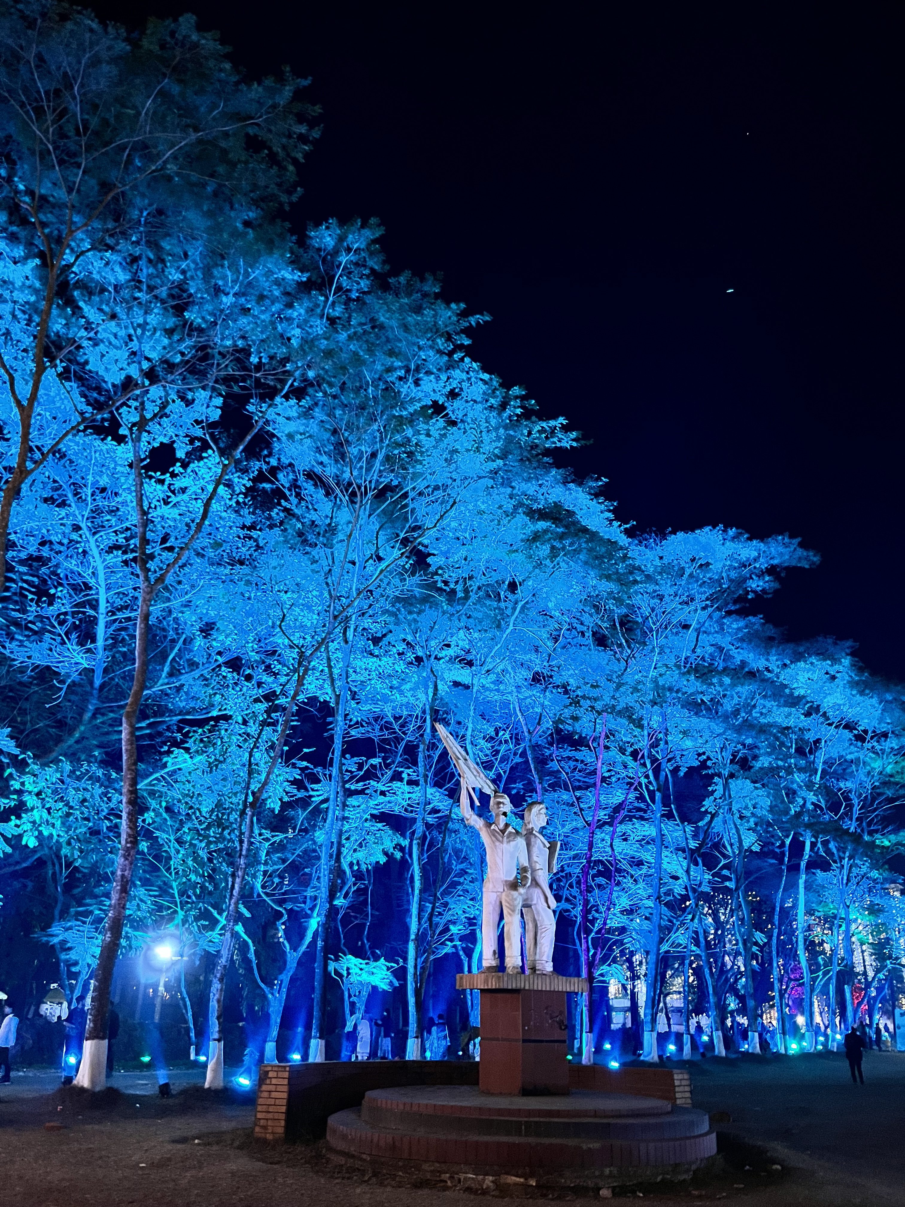A statue of two figures holding a flag stands on a circular pedestal surrounded by trees illuminated with blue lights at night. The background features more illuminated trees and faint silhouettes of people.