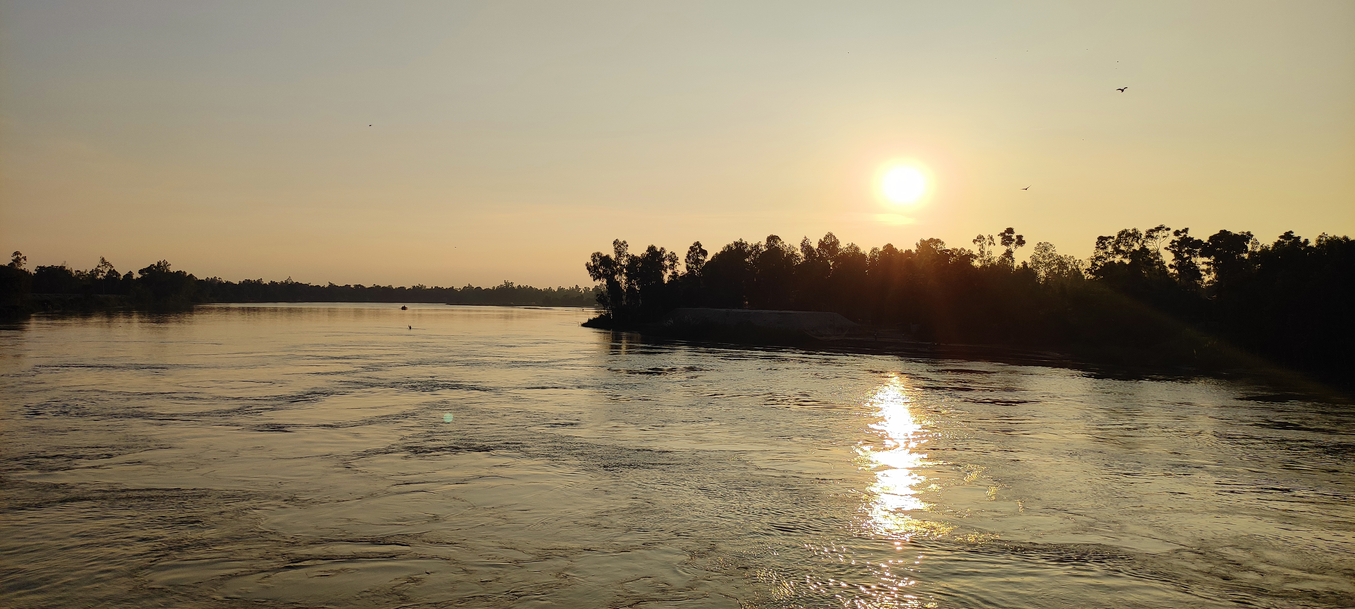 A serene river scene at sunset, featuring calm waters reflecting the orange and yellow hues of the setting sun.