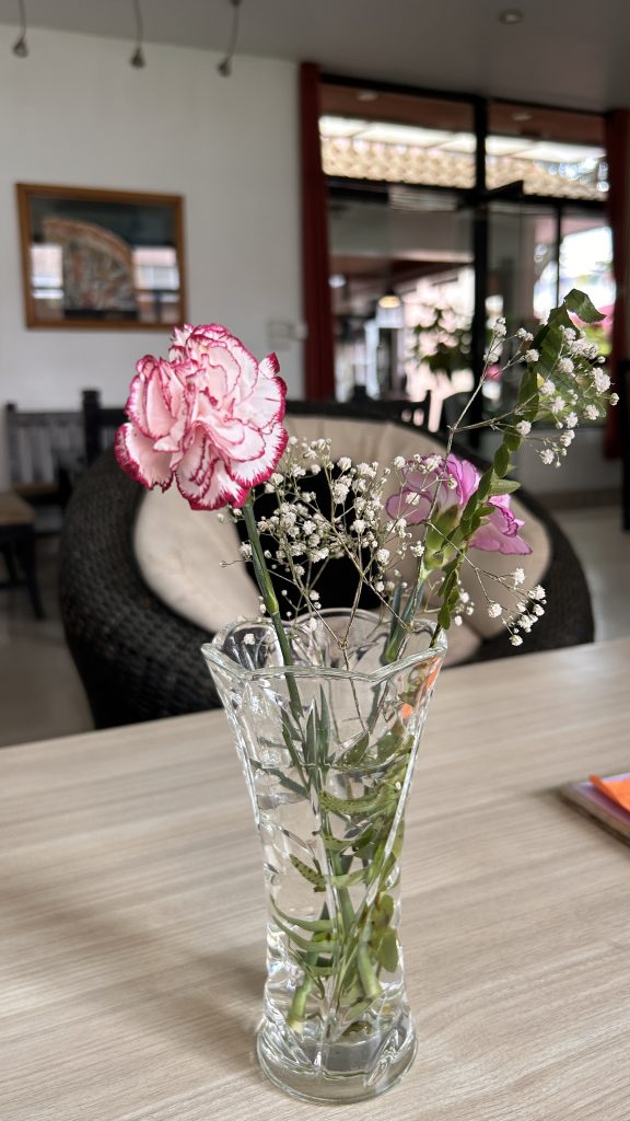 A clear glass vase filled with water holds several flowers, including a pink and white carnation, small white baby’s breath flowers, and a few purple blossoms