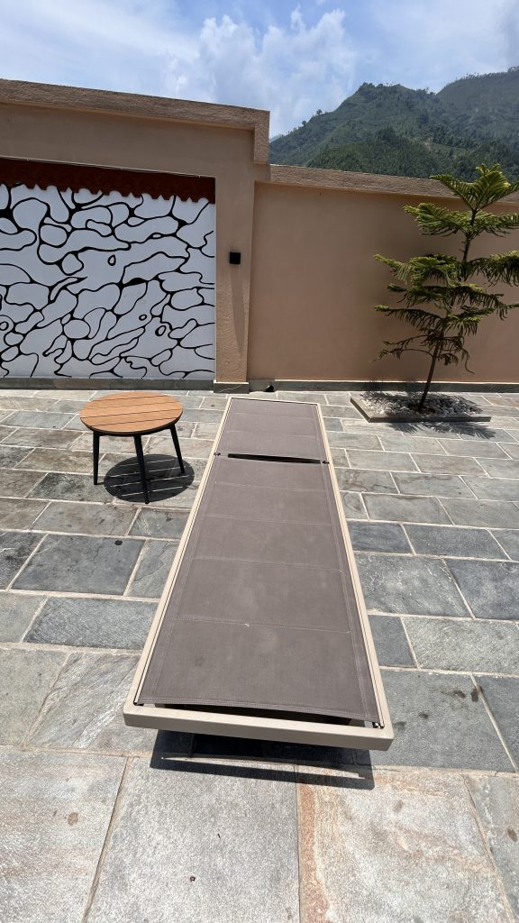 A patio area featuring a stone floor, a low round wooden table with black legs, and a long, gray lounge chair positioned horizontally.