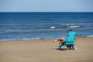 A person relaxes in a blue beach chair on a sandy beach, facing the ocean. They have bare feet resting on the sand and are enjoying the view of the calm blue water and gentle waves.