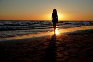 A silhouette of a person walking along a beach at sunset. The scene features gentle waves lapping at the shore, with the sun setting just above the horizon, casting warm hues of orange and yellow across the sky and reflecting on the wet sand.