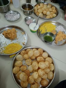 A casual spread of snacks features crispy puris, creamy yogurt, sev, chutneys, and spiced broth arranged on metal plates.