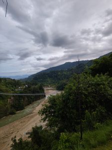 A nature scenario with cloudy sky, green hills, jungles and suspension bridge. A river is flowing down across the bridge.