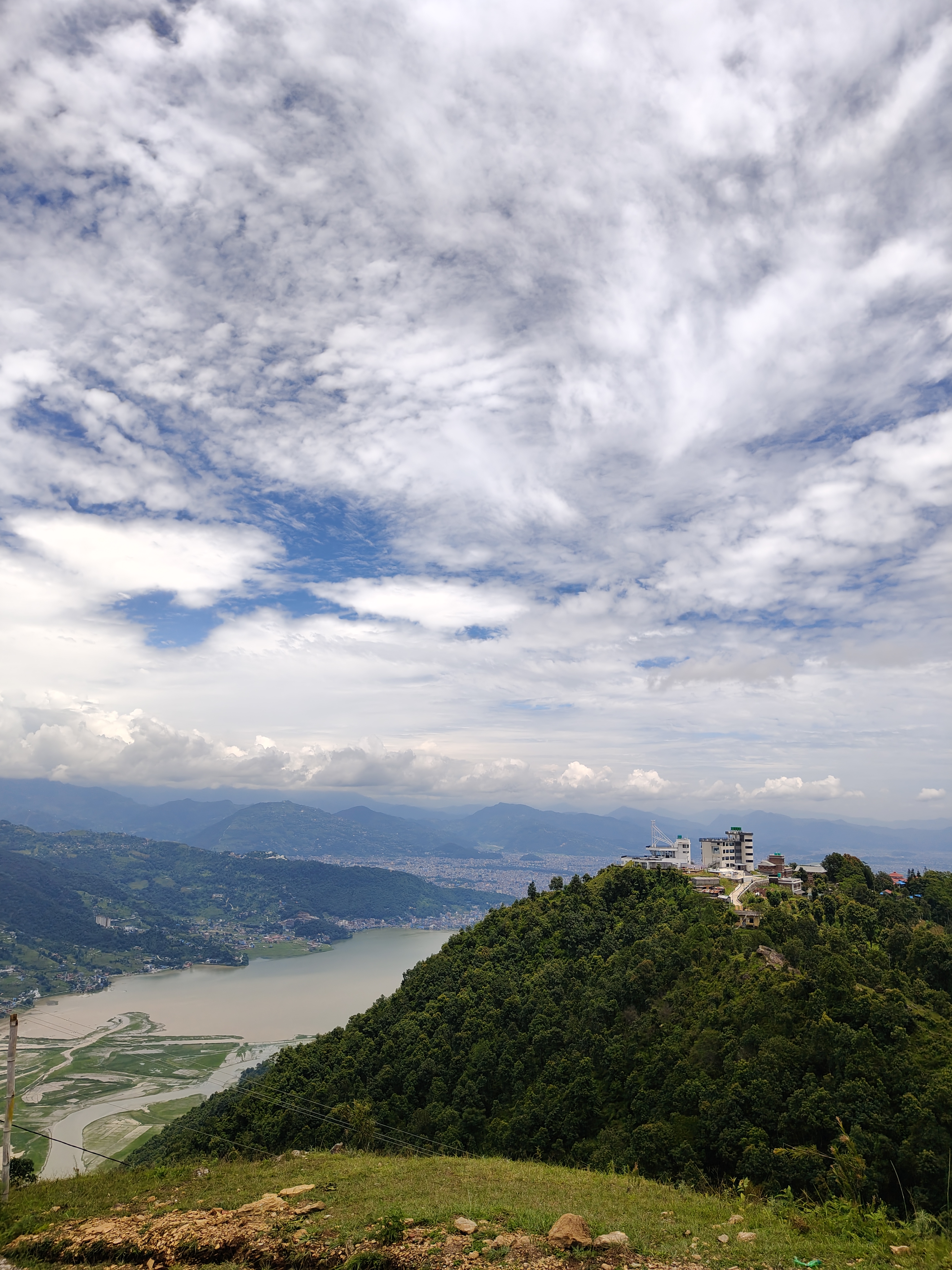 A scenic landscape featuring a lush green hill rising above a lake, with a small cluster of buildings at its peak. In the background, a valley surrounded by mountains is visible, while the sky above is partly cloudy with patches of blue.