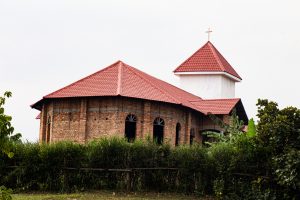 A brown brick church with a red tiled roof, featuring a white section with a small cross on top.
