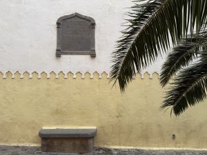 A basalt plaque set into a two-tone ochre-and-white plaster wall marks Captain Juan Rejón’s founding of Las Palmas on the night of San Juan in 1478; beneath it, a simple stone bench sits on the cobblestones, while palm fronds arch into the frame from the right.
