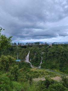 A scenic landscape featuring lush green hills with waterfalls cascading down one side. In the foreground, dense vegetation includes trees and shrubs. A river runs along the base of the hills, winding through the valley. 