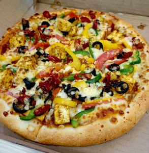 Close-up of a loaded vegetarian pizza topped with paneer cubes, bell peppers, black olives, red chilies, and melted cheese inside a cardboard box.