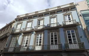 Elegant light blue European building with white trim, 10 large windows, and decorative wrought iron balconies.