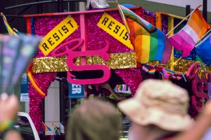 A colorful Pride parade float decorated with glitter, gold fringe, and large pink lettering features signs reading “RESIST.” Progress Pride, lesbian, and other LGBTQ+ flags wave from the top, while spectators cheer and wave fans in the foreground. #shareyourpride2025