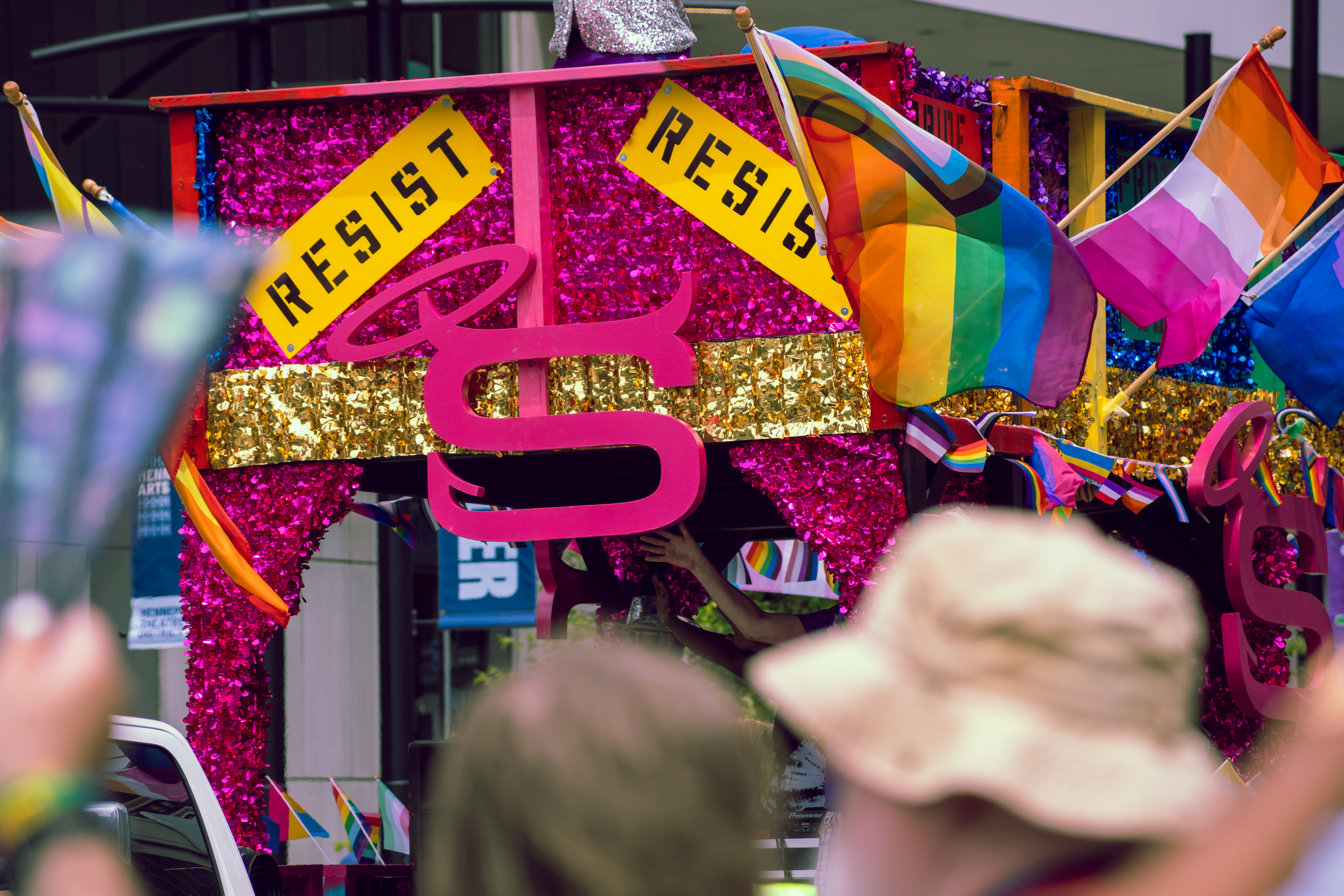 A colorful Pride parade float decorated with glitter, gold fringe, and large pink lettering features signs reading “RESIST.” Progress Pride, lesbian, and other LGBTQ+ flags wave from the top, while spectators cheer and wave fans in the foreground. #shareyourpride2025